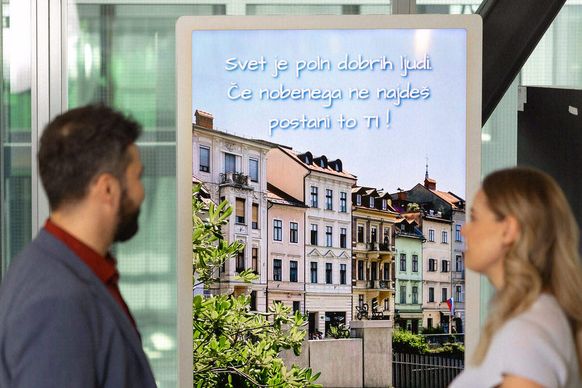 Dve osebi stojita pred digitalnim zaslonom, na katerem je prikazana slikovita podoba pisanih stavb in klopi z besedilom v tujem jeziku.