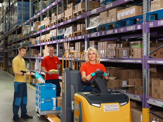 Sodelavci v logističnem centru organizirajo pakete in pripravljajo blago za skladiščenje ter oskrbo dm prodajaln. Ena oseba upravlja viličar, drugi sodelavci pa skrbijo za razvrščanje izdelkov, upravljanje zalog in nemoten potek logističnih procesov v skladišču.