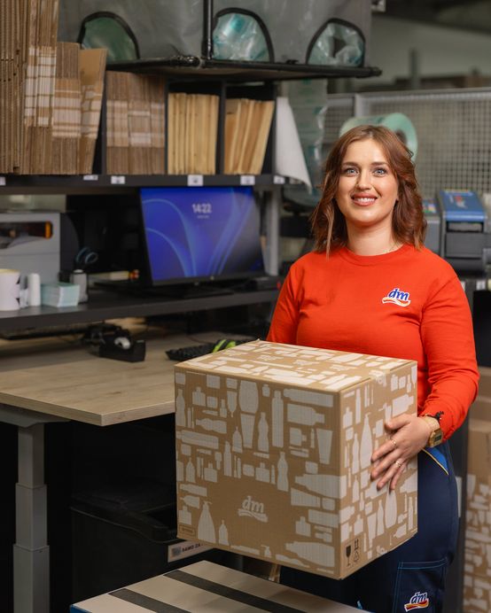 Sodelavka v logističnem centru drži kartonsko škatlo ob delovni postaji. V ozadju so skladiščne police, računalnik in materiali za pakiranje, ki podpirajo pripravo blaga, logistične procese in oskrbo dm prodajaln.