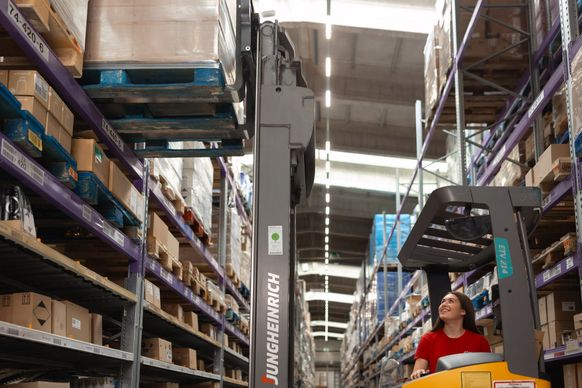 Sodelavka v logističnem centru upravlja viličar in dviguje paleto na visoko skladiščno polico v skladiščnem hodniku.