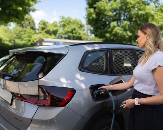 Ženska priklaplja električno vozilo na polnilno postajo v sončnem zunanjem okolju,