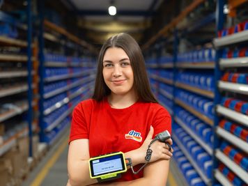 Sodelavka v logističnem centru stoji med skladiščnimi regali in uporablja zapestni čitalnik črtnih kod.