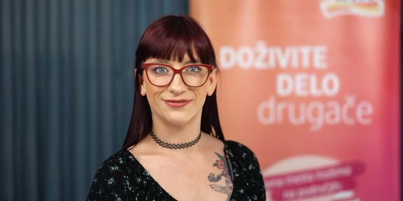 Nasmejana ženska z rdečimi očali in tetovažo na prsnem košu stoji pred tablo z besedilom v slovenščini ter gleda v kamero.
