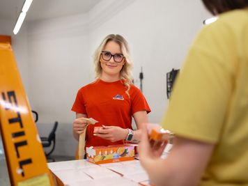 Sodelavka v rdeči majici in očalih lepi etikete na škatlo v logističnem centru, medtem ko sodeluje z drugo osebo.