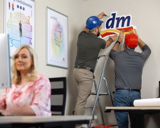 Dva delavca z zaščitnima čeladama nameščata logotip »dm« na steno, medtem ko ženska v ospredju za pisalno mizo uporablja računalnik.