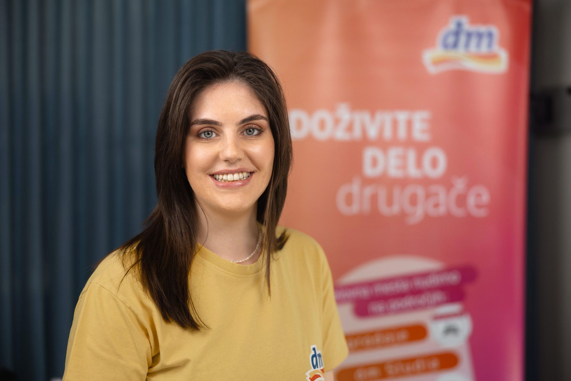 Nasmejana oseba v rumeni majici stoji pred barvitim ozadjem z besedilom in logotipom blagovne znamke.
