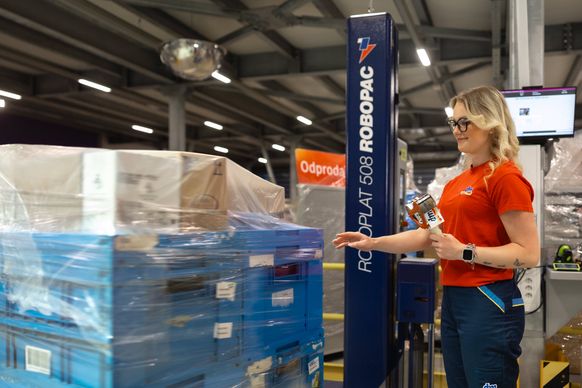 Ženska v rdeči majici upravlja ovijalni stroj v logističnem centru in pripravlja paleto, ovito v zaščitno folijo.