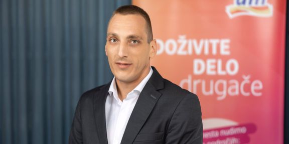 Moški v poslovni obleki stoji pred oranžnim panojem z besedilom in logotipom dm ter pozira za portretno fotografijo.