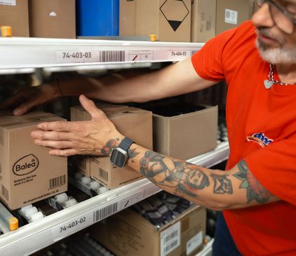 Oseba v rdeči majici in s pametno uro razvršča izdelke Balea na skladiščno polico v logističnem centru. Na rokah ima tetovaže.