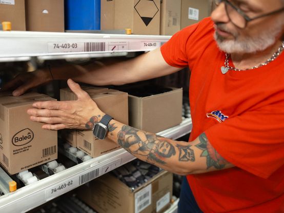 Oseba v rdeči majici in s pametno uro razvršča izdelke Balea na skladiščno polico v logističnem centru. Na rokah ima tetovaže.