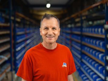 Sodelavec v logističnem centru stoji med skladiščnimi regali z zaboji in izdelki. V okviru logističnih procesov skrbi za upravljanje zalog, organizacijo blaga ter nemoteno oskrbo dm prodajaln.