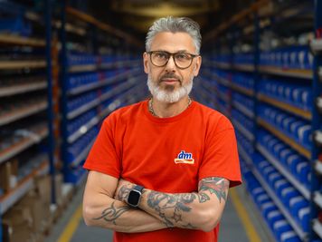 Oseba s tetovažami, v rdeči majici in očali stoji s prekrižanimi rokami med vrstami modrih skladiščnih regalov.