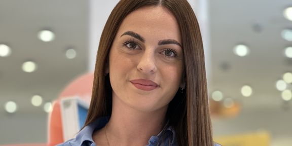 Nasmejana zaposlena v dm z dolgimi rjavimi lasmi, oblečena v modro polo majico z imensko priponko, stoji v sodobni dm prodajalni
