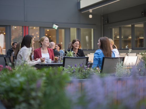 Eine Gruppe von Mitarbeitenden sitzt im Innenhof eines Büros und genießt Getränke und Snacks. Umgeben von Pflanzen und einer einladenden Atmosphäre, führen sie Gespräche.